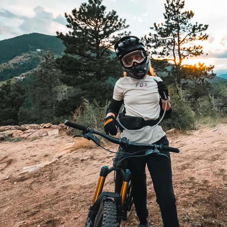 A mountain biker wearing a black helmet, goggles, and a white Fox jersey stands on a dirt trail with a mountain bike in front of a scenic forested mountain landscape at sunset. She holds a drinking tube in her left hand.