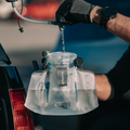 Person filling a HydraPak 1.5 litre Contour lumbar reservoir from a bulk reservoir using the Camp Tap. Reservoir is clear with blue markings for measurements.