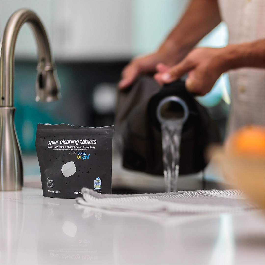 Packaging of Bottle Bright gear cleaning tablets on a kitchen counter with a person washing a water reservoir in the background.