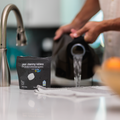 Packaging of Bottle Bright gear cleaning tablets on a kitchen counter with a person washing a water reservoir in the background.